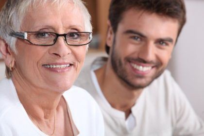 And older woman with white hair and a young man with brown hair sit near each other, looking at the camera and smiling.