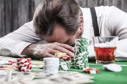 A distraught gambler lays his head down on his hands at a poker table, with stacks of chips and a drink on the table in front of him.