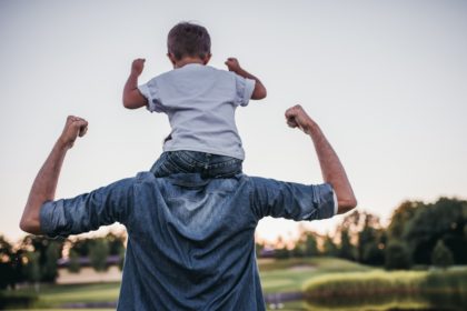 A small child sits on his father's shoulders while they both flex their muscles