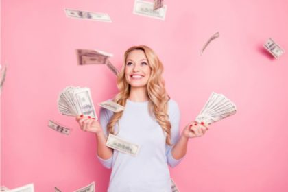 A blond woman in front of a pink wall is holding two handfuls of hundred dollar bills, smiling while money flutters down all around her.