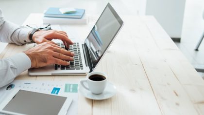 man using a laptop on a wooden desk with coffee and paperwork