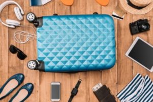 A teal hard suitcase surrounded by accessories like shoes, a hat, a camera, a wallet, etc.