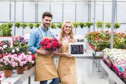 A man and woman in a flower shop. The man holds a bouquet of red flowers and the woman holds a chalkboard "OPEN" sign.