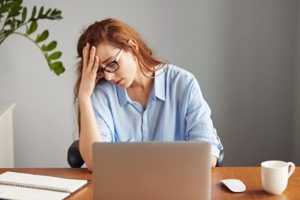 A distressed woman with red hair sits in front of a white laptop, leaning her head on one hand.