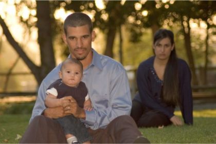 A father sits in the forefront of the photo, holding a baby in his lap while the mother sites in the background, looking upset.
