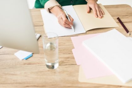 A woman writes in a file at a desk covered in papers and a glass of water.