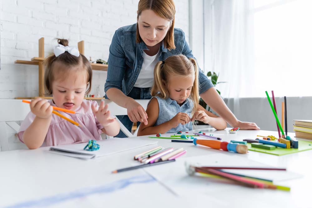 two preschool girls working at a table in preschool while their teacher points at something on the table from over their shoulders - child support and special needs children