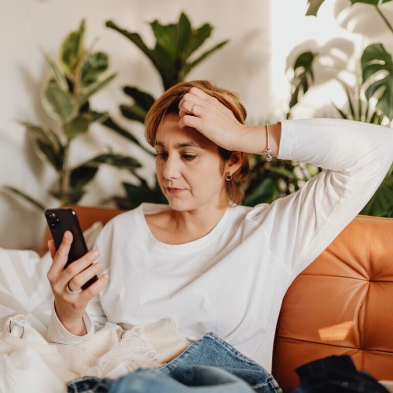 Woman sitting on couch looking stressed with her hand on her head as she looks at her phone.