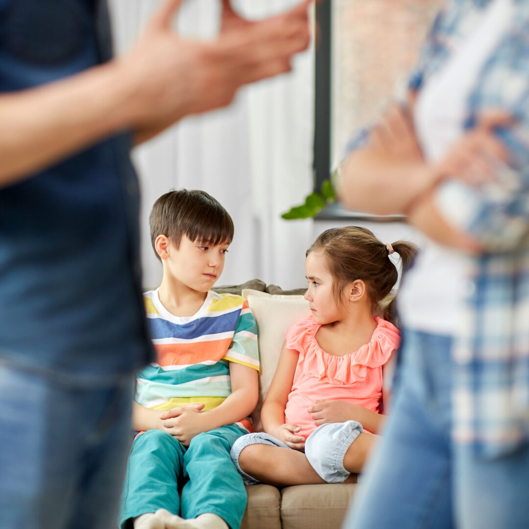 Cute girl and boy 4-5 years old sitting on couch and looking at each other with upset looks while faded image of parents are arguing
