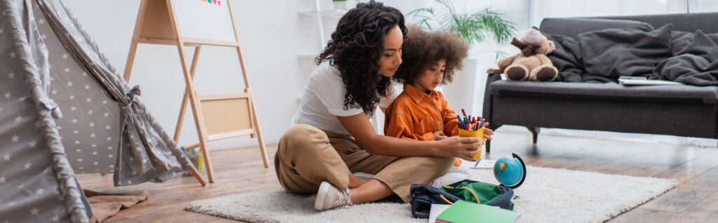 Young african american woman holding colorful pencils near backpack, globe and tent at home, banner,stock image