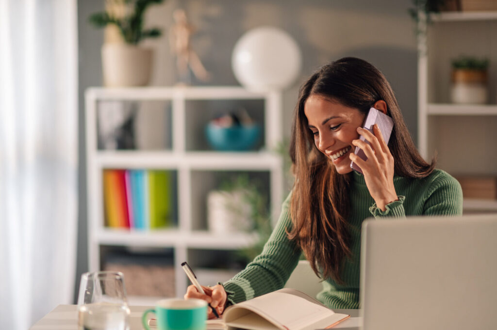 Young businesswoman working at home while talking on a phone and taking notes. Home remote work. Beautiful female talking on a smartphone with her client and writing his ideas down. Copy space.
